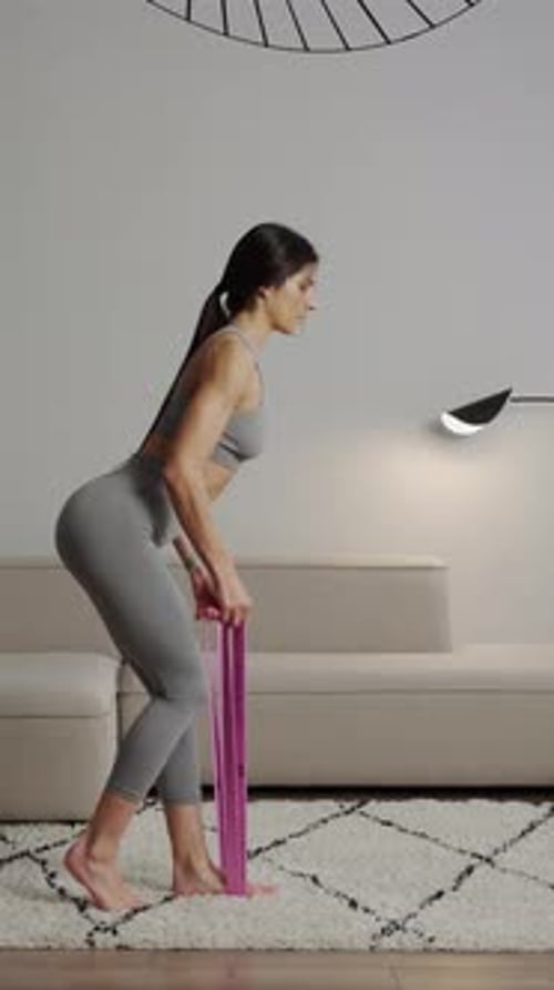 Athletic Woman in Gray Sportswear Performing Resistance Band Deadlift Exercise at Home on Soft Rug