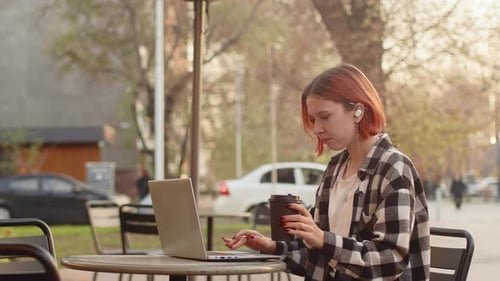 Young Caucasian Woman in Casual Clothes Drinking Coffee From a Paper Cup and Working on a Laptop