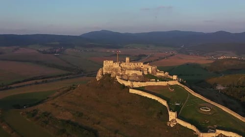 A drone flies toward the illuminated Spiš Castle during sunset, revealing its stone towers, golden g