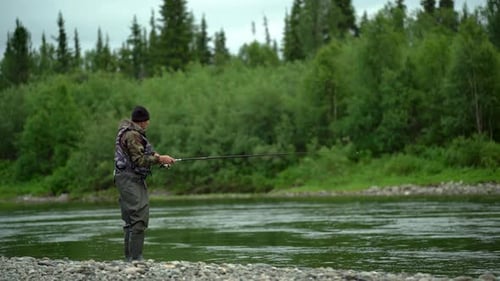 A Fisherman Stands On The River Bank And Catches Fish With A Spinning Rod Near The Forest