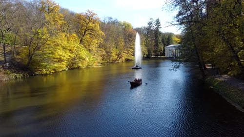 People Sailing in Boat on Lake Decorative Fountain Trees Yellow Leaves in Park