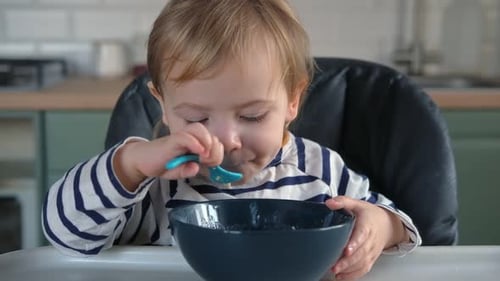 One Year Old Hungry Baby Girl in Striped Casual Clothes Sits at White Table in Highchair and Eats