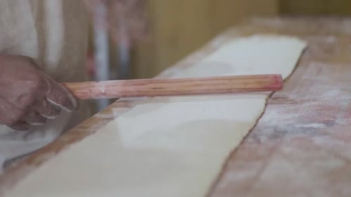 Close-up of a pastry chef rolling dough with hands using a wooden rolling pin on a work surface.