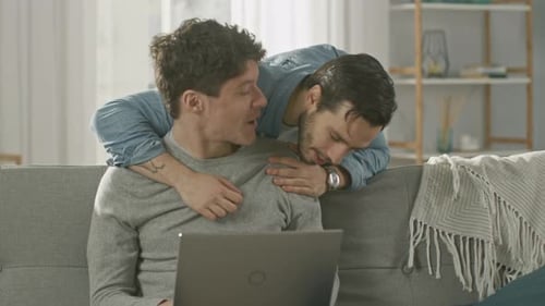 Cute Male Gay Couple Spend Time at Home. Young Man Works on a Laptop, His Partner Comes From Behind