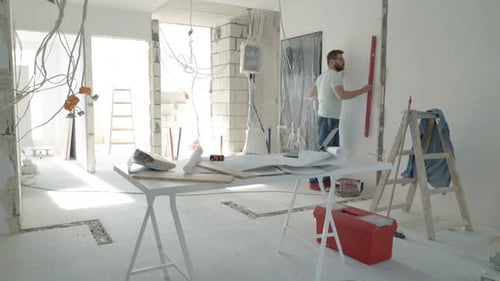 Männlicher Architekt mit Bauplänen überprüft Wand mit Ebene im neuen Zuhause