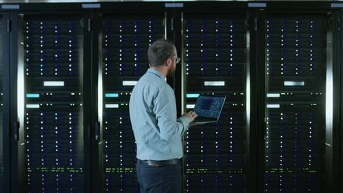 Bearded IT Specialist in Glasses is Working on Laptop in Data Center while Standing Before Server R