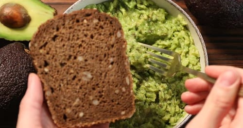 Woman spreading avocado dip onto slice of bread at table, top view