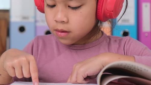 Fröhliches kleines Mädchen mit Kopfhörern, das beim Lesen eines Buches singt, sitzt am Tisch in ihrem Zimmer