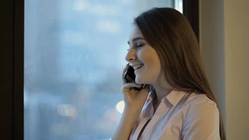 Young woman chatting on cellphone by window at home in the city at evening