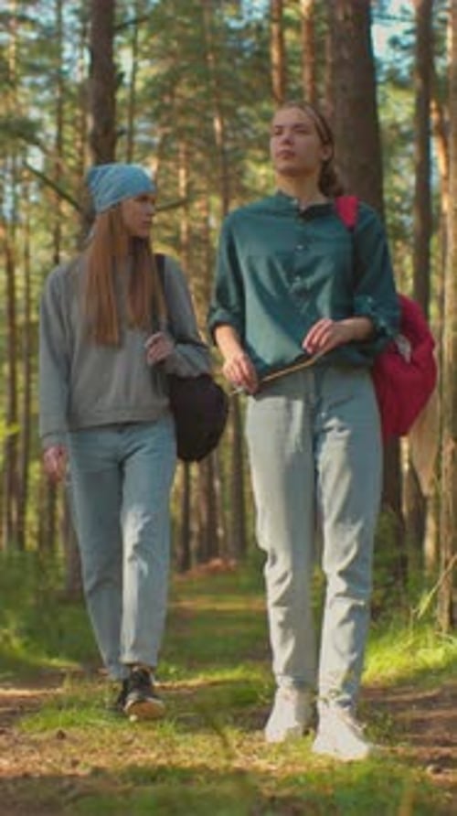 Hikers Walking Through Forest One with Blue Scarf Gazing Into Trees Other Holding Grass