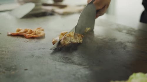 Chef Slicing Chicken Meat Using Knife With Shrimps On Griddle Pan. - close up