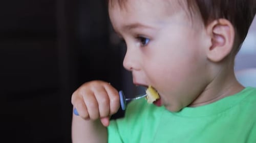 Cute baby boy wearing green t-shirt eating from fork. Mom helps her son to pick up the food.