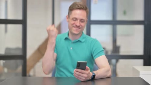 Young Man Celebrates Success Using Smartphone in Office