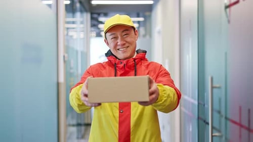 Junger lächelnder Zusteller macht eine Schenkgeste mit Kartonschachtel im Innenbereich