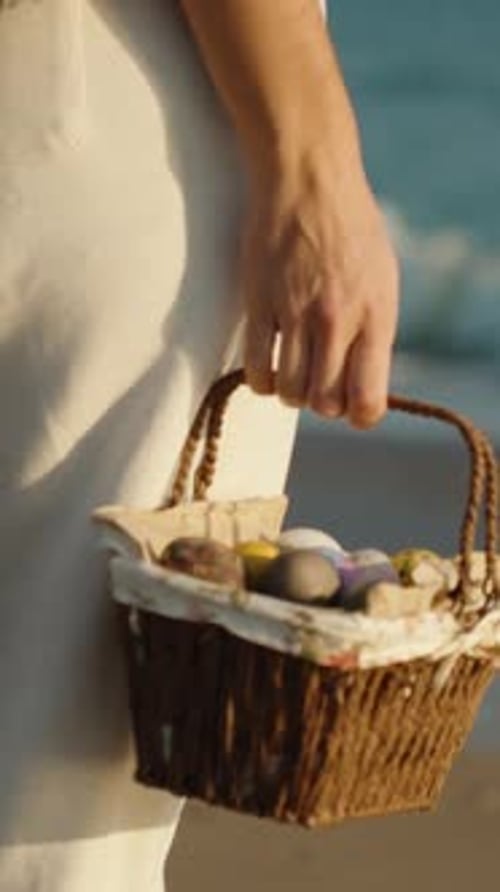 A woman with a basket of Easter Eggs walks on the sand, stops and looks into the sea. Vertical video