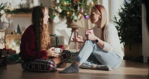Hermosas hermanas con tazas de café hablando en el suelo de casa durante Navidad