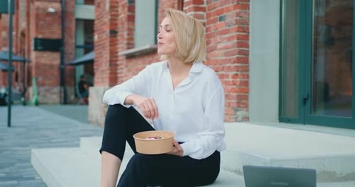 Woman eating healthy salad with olive from plastic container sitting on stairs in city street, sunli