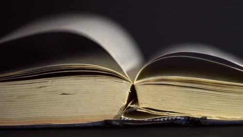 Slow motion, close up of an old, vintage, open book with pages turning on the wind, on a dark room w
