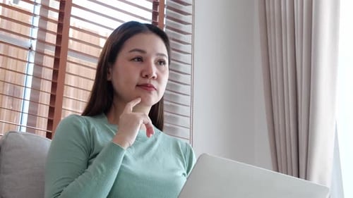 Young asian business woman working with laptop while thoughtful sitting on sofa in home office.