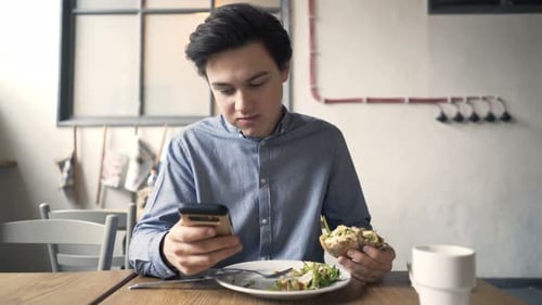 Teenager With Smartphone Eating Hamburger Sitting In Cafe