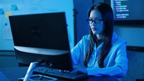 Konzentrierte asiatische Softwareingenieurin mit Brille, programmiert spät in der Nacht auf einem Desktop-Computer