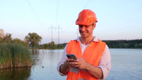 Happy Male Engineer in Safety Helmet Typing on the Smartphone Highest Voltage Transmission River on