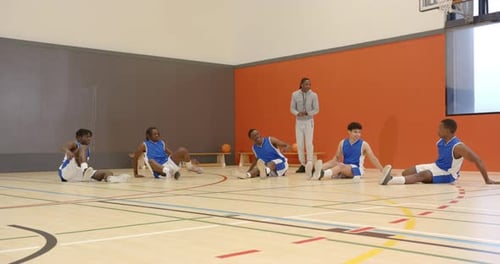 Basketball players stretching on court with coach supervising training session