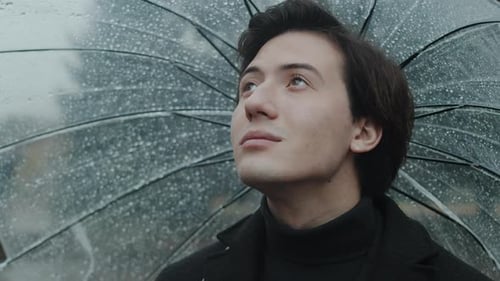 Young Man in a Coat Standing Under a Wet Transparent Umbrella in the City Park Close Up Looking Up