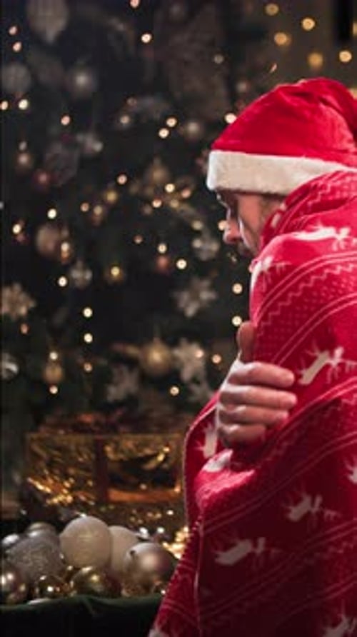 Cold Room Man in New Year's Hat and Raincoat Warms Himself Near Christmas Tree in Evening at Home