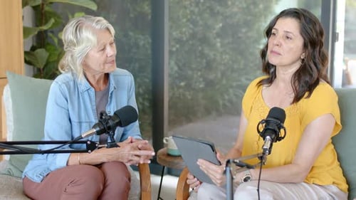 Female hosts hosting podcast with mics, tablet causing right host reading, gesturing, left nodding