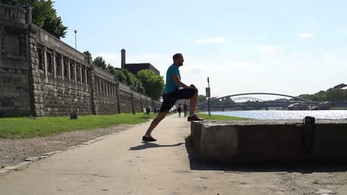 Man jogger stretching legs by river in city park preparing for workout in slow motion