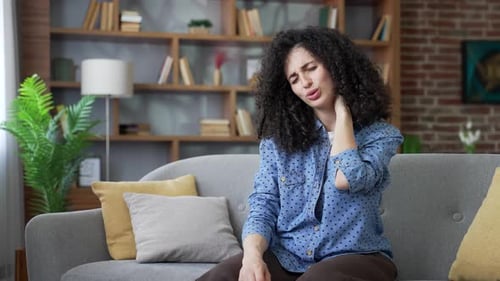 Tired young woman suffering from neck pain while sitting on sofa in living room at home.