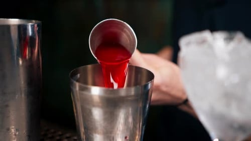 Close Up Male Bartender Masterfully Pours Beverage Into Steel Jigger Next to It is Tall Transparent