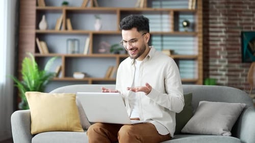 Young adult man talking on a video using a laptop sitting on sofa in living room in home office.