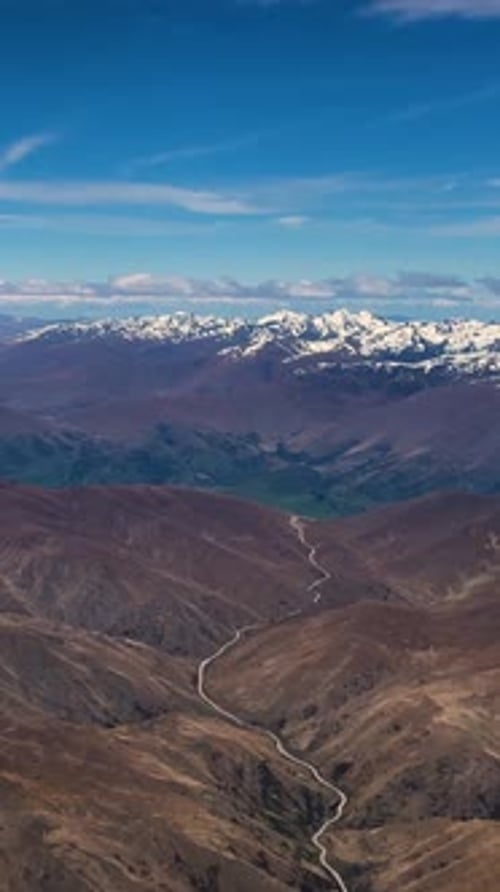 Aerial of road winding through brown mountains with The Remarkables in the distance