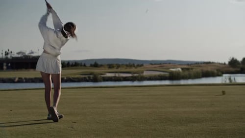 Young Woman Golfer Swings Club to Hit Ball on Green Lake Bank
