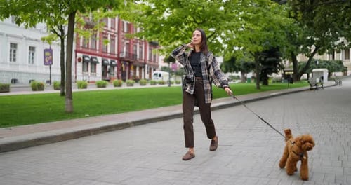 A Happy Brunette Girl in a Plaid Shirt Walks with Her Curly Brown Dog Along the Street in the City