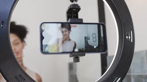 African American woman recording after red dot on phone in bathroom ring light demoing skincare