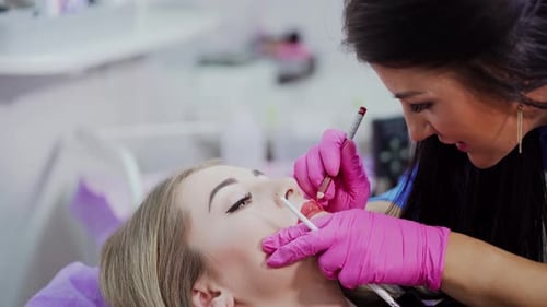 Cosmetic procedure of tattoo on lips. Master applying red permanent makeup on young girl lips.