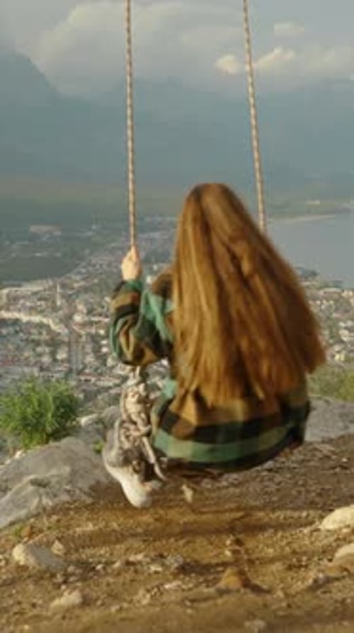 Vertical video. At the mountain summit, near the edge of the cliff, a young woman fearlessly swings