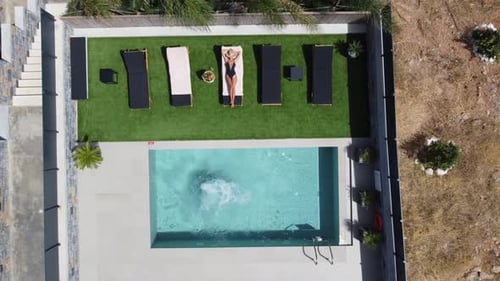 Drone flies over an outdoor pool in bird's eye view - Woman in swimming costume lies on sunbed and m