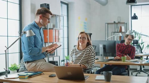 Male Colleague Sits on a Table in Front of a Young Female Cowroker while Holding a Tablet Computer