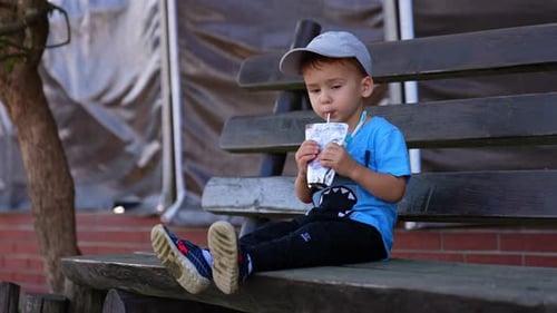 Calm Caucasian baby boy sitting on the bench outdoors drinking juice from doy pack. Thirsty child dr
