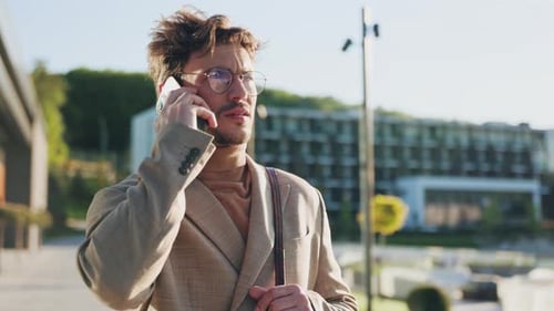 Young Man with Glasses Talking on a Mobile Phone in a City in the Sunlight Confident Young