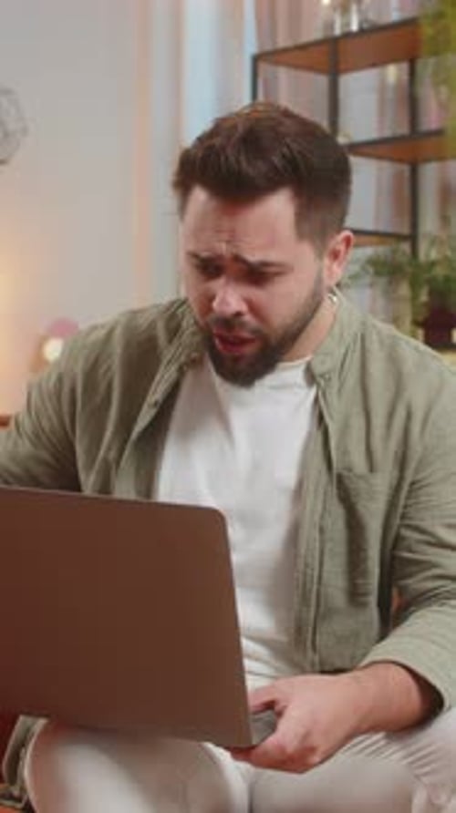 Un joven caucásico molesto que usa una computadora portátil entabla acaloradas conversaciones sentado en el sofá