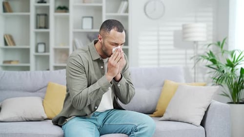 Man Sneezes into Tissue on Couch in Living Room