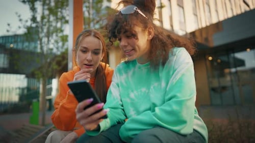 Two Young And Stylish Female Students Talking On City Street And Using Smartphone To Scroll Social