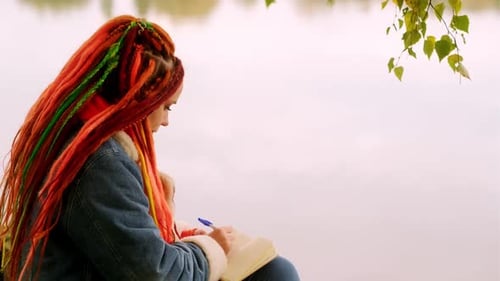 Young Creative Woman with Dreadlocks Drawing Writing in Notebook Sitting on Stumps Near Lake in