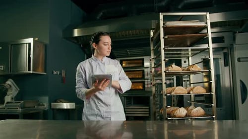 Female Baker Reviews Inventory on Tablet in Kitchen