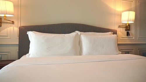 Luxury accommodations with large soft bed, fluffy pillows and lamp shining on wall of hotel suite. C